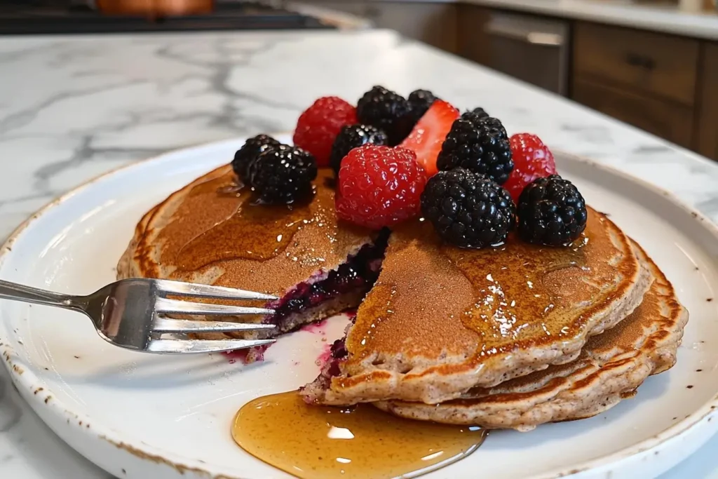 A fork cutting into a stack of buckwheat pancake mix pancakes with honey and fresh berries.