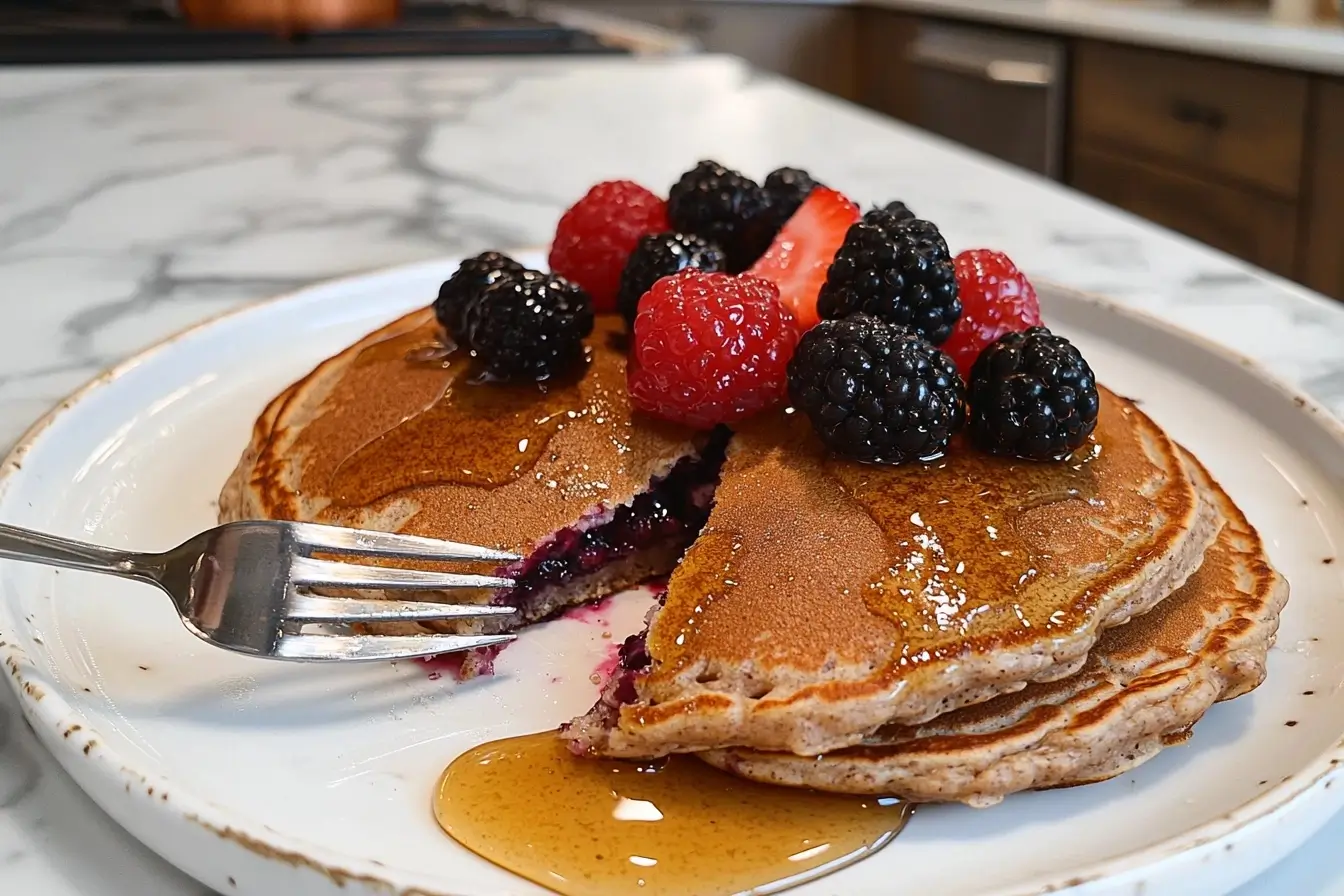 A fork cutting into a stack of buckwheat pancake mix pancakes with honey and fresh berries.