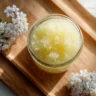 Homemade sugar scrub in a jar with ingredients like sugar and oil on a wooden surface.
