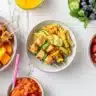 Overhead view of colorful, healthy and kid-friendly meal components for picky eaters, including chicken skewers, hidden veggie pasta, and a smoothie, on a marble counter.