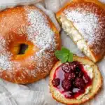 Overhead view of two golden vegan cottage cheese bagels, one sliced with berries and mint, on a light wooden surface